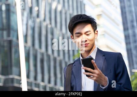 Junger asiatischer Geschäftsmann, der beim Gehen Nachrichten auf dem Handy ansieht In der Straße in der Innenstadt der modernen Stadt Stockfoto