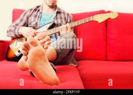 Barfuß-Mann im karierten karierten karierten Hemd sitzt auf dem roten Sofa am Fenster und spielt Offset-E-Gitarre, Mahagoni-Hals. Rockgitarrist spielt Jazz impro Stockfoto