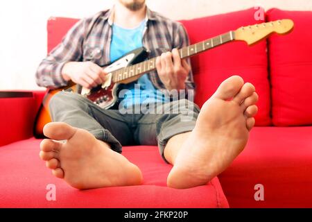 Barfuß-Mann im karierten karierten karierten Hemd sitzt auf dem roten Sofa am Fenster und spielt Offset-E-Gitarre, Mahagoni-Hals. Rockgitarrist spielt Jazz impro Stockfoto