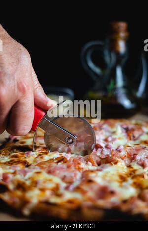 Ein Mann, der mit einem Pizzaschneider eine Pizza schneidet. Im Hintergrund stehen Oregano und Olivenöl. (Vertikal) Stockfoto