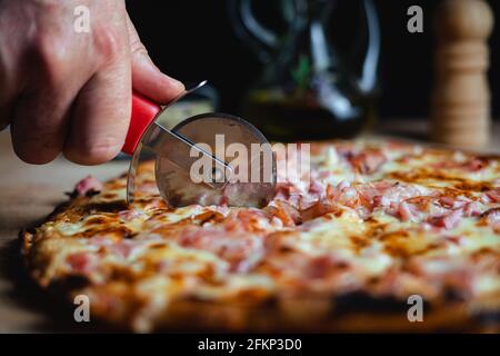 Ein Mann, der mit einem Pizzaschneider eine Pizza schneidet. Im Hintergrund stehen der Orangeano, das Olivenöl und der Pfeffer. (Horizontal) Stockfoto