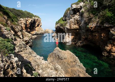 Ein Mann in roten Badehosen, der auf einem Felsen steht, bereitet sich darauf vor, in das kristallklare Wasser von cala rafalet auf menorca, spanien, zu tauchen Stockfoto