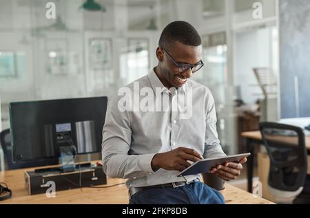 Lächelnder afrikanischer Geschäftsmann, der sich mit einem an einen Schreibtisch lehnt Tablet Stockfoto