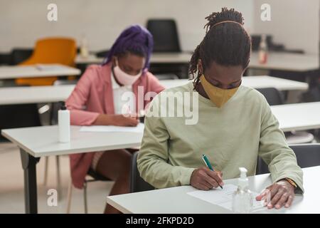 Porträt eines jungen afroamerikanischen Mannes, der während der Prüfung oder Prüfung in der Schule eine Maske trägt, mit Händedesinfektionsmittel, Kopierraum Stockfoto