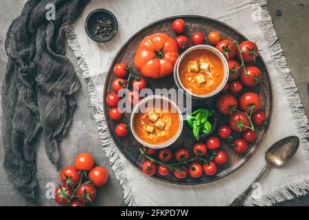 Zwei Tassen gefüllt mit frischer, hausgemachter Tomatensuppe auf einem Holztablett mit einer Vielzahl von reifen Tomaten, Draufsicht Stockfoto
