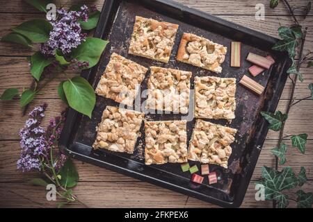 Backblech mit Rhabarber-Krümelkuchen auf Holzboden, mit Fliederblüten verziert, Draufsicht Stockfoto