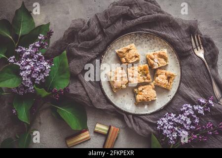 Stücke von Rhabarber zerbröseln Kuchen auf einem Teller auf einem grauen Hintergrund, dekoriert mit Fliederblumen, Draufsicht Stockfoto
