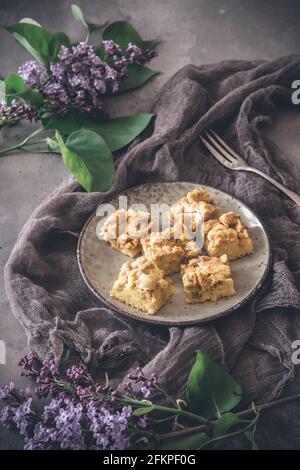 Stücke von Rhabarber zerbröseln Kuchen auf einem Teller auf einem grauen Hintergrund, mit Fliederblumen verziert, vertikal Stockfoto