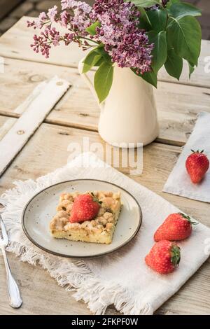 Ein Stück Rhabarber-Krümel-Kuchen und Erdbeeren auf einem Teller auf einem alten Holztisch, verziert mit Fliederblumen, vertikal Stockfoto