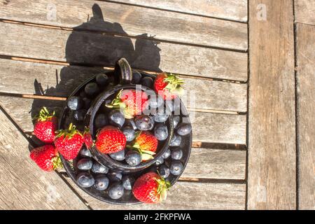 Eine flache Komposition aus einer Teetasche, die bis zum Überlaufen mit Erdbeeren und Heidelbeeren auf einer verwitterten, hölzernen Oberfläche gefüllt ist Stockfoto