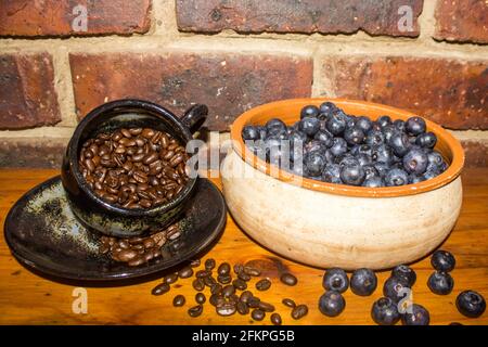 Ein Stillleben, bestehend aus einer Schüssel voller Heidelbeeren und einer Tasse gefüllt mit Kaffeebohnen vor einem Backstein Hintergrund Stockfoto