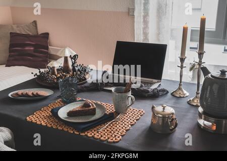 Tisch mit Laptop, Kuchen, Keksen und herbstlicher Dekoration in einem gemütlichen Raum für virtuellen Kaffee mit Familie oder Freunden vorbereitet, nützlich während der weltweiten CO Stockfoto
