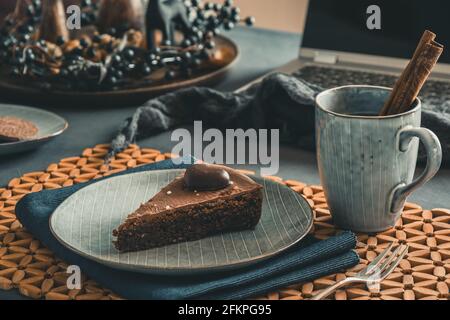 Stück hausgemachte Schokoladenkuchen und eine Tasse mit Tee und Zimtstange auf einem grauen Teller auf einem herbstlich dekorierten Tisch, Laptop im Hintergrund öffnen. Stockfoto