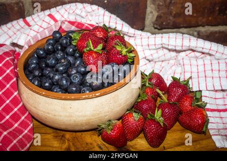 Eine rustikale Töpferschale, gefüllt mit Heidelbeeren, umgeben von Erdbeeren und roten Küchentüchern Stockfoto