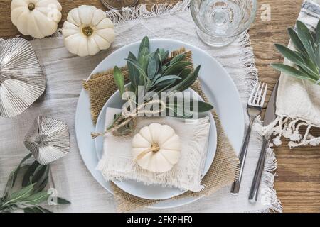 Draufsicht der Platzeinstellung auf einem Holztisch mit weißen Mini-Kürbissen, Salbeiblättern und Kristallgläsern für Thanksgiving Day oder Halloween Stockfoto