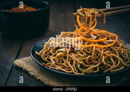 Essstäbchen nehmen chinesische vegetarische Nudeln, serviert auf schwarzem Teller auf dunklem Holzgrund Stockfoto