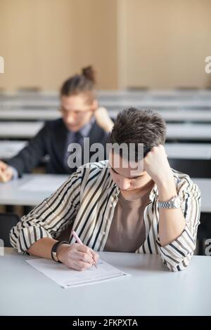 Vertikales Porträt eines jungen Mannes, der in der Schule eine Prüfung macht, während er im Auditorium am Schreibtisch sitzt und denkt, Raum kopiert Stockfoto