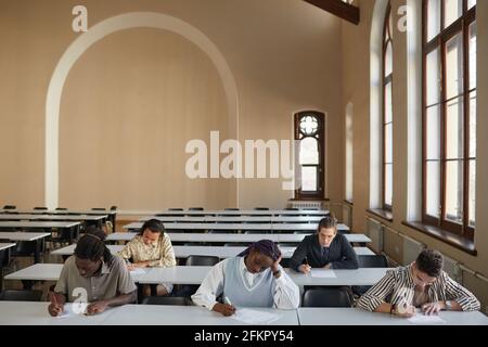 Weitwinkelansicht auf verschiedene Gruppen von Studenten, die die Prüfung ablegen, während sie am Schreibtisch im Schulsaal sitzen, Kopierfläche Stockfoto