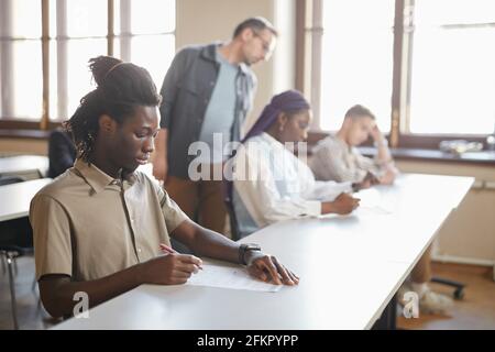 Eine vielfältige Gruppe von Studenten, die am College die Prüfung ablegen, während sie in Reihe am Schreibtisch im Auditorium sitzen, konzentriert sich auf den jungen afroamerikanischen Mann im Vordergrund, Kopie s Stockfoto