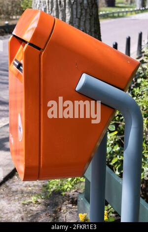 Typischer holländischer orangefarbener Briefkasten mit PostNL-Logo, der sich im Frühling in einer Straße entlang eines Parks mit Bäumen und Sonne befindet. Vertikales Bild, Seitenansicht Stockfoto