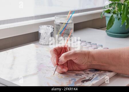 Die Hand der älteren Frau hält einen Pinsel. Sie malt mit Acrylfarben nach Zahlen auf Leinwand. Digitales Detox-Konzept Stockfoto