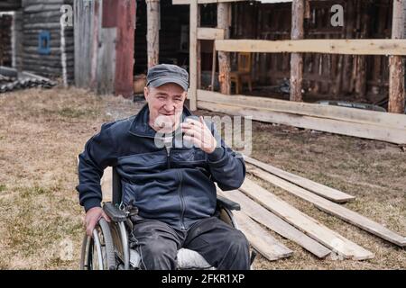 Porträt eines glücklichen älteren Mannes in legerer Kleidung in einem Rollstuhl im Hof seines Hauses, der lächelt und die Kamera anschaut. Eine Zigarette in der Hand. Stockfoto