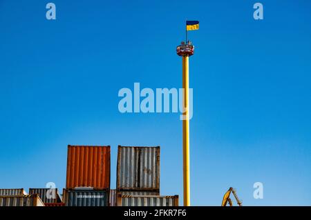 Container-Terminal im Frachthafen der Ukraine. Hoher Fahnenmast mit wehender ukrainischer Flagge auf klarem blauen Himmel Hintergrund in der Nähe von alten gestapelten Containern mit s Stockfoto
