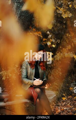 Nachdenkliche rothaarige Frau im Park auf einer Holzbank. Hübsche Frau in Baskenmütze und Lederjacke. Eine Person mit einem Grafiktablett, ein Designer, ein Freiberufler. Herbstporträt. Stockfoto