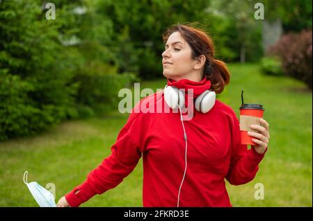 Eine rothaarige Frau mit Kopfhörern trinkt im Park Kaffee mit Stroh und zieht eine medizinische Maske aus. Das Mädchen genießt einen freien Spaziergang in einem sonnigen Sommer Stockfoto