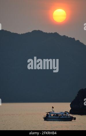 Panoramablick auf den Sonnenuntergang über dem glitzernden Wasser, den Felsformationen, den blauen Bergen und den schwimmenden Booten in der Halong-Bucht in Vietnam Stockfoto