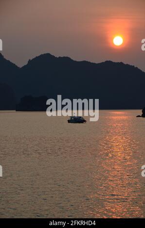 Panoramablick auf den Sonnenuntergang über dem glitzernden Wasser, den Felsformationen, den blauen Bergen und den schwimmenden Booten in der Halong-Bucht in Vietnam Stockfoto