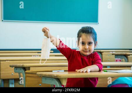 Das kleine Mädchen zieht ihre schützende Gesichtsmaske aus und hebt sich Ihre Hand im leeren Klassenzimmer Stockfoto