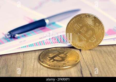 Eine Münze der Bitcoin-Kryptowährung steht an ihrem eigenen Ende, die zweite auf den Diagrammen der Änderungen der Kryptowährungsraten auf einem Holztisch Stockfoto