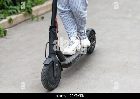 Die Beine eines unbekannten Mädchens in weißen Sneakers reisen auf einem schwarzen Elektroroller auf einem dunklen städtischen Asphalt. Stockfoto