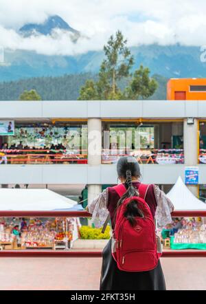 Junge indigene Otavalo-Frau mit Schulrucksack und traditioneller Kleidung auf dem modernen lokalen Markt mit dem Vulkan Imbabura im Hintergrund, Ecuador. Stockfoto