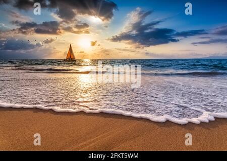 Ein Segelboot segeln entlang des Ozeans gegen EINE bunte Ocean Sunset Sky als sanfte Welle Rolls to Shore Stockfoto