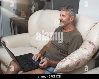 Lächelnder Mann mittleren Alters, der mit dem auf der Couch sitzt Laptop online zu Hause kaufen Stockfoto