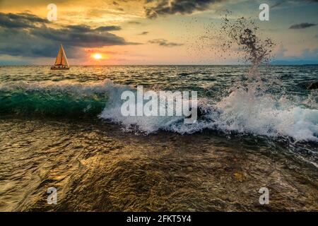 Ein Segelboot segeln entlang des Ozeans gegen EINE bunte Ocean Sunset Sky als sanfte Welle Rolls to Shore Stockfoto