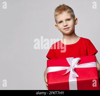 Porträt eines lächelnden Jungen im roten T-Shirt mit großem Geschenkkarton mit Band in den Händen, es zu tragen, bereit für den Geburtstag Stockfoto