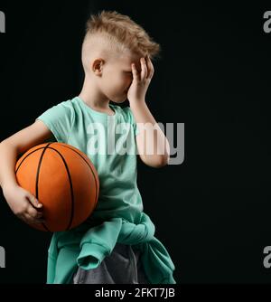 Blonde junge in T-Shirt, Hose und Hoodie um die Taille steht hält Basketball und Gesten facepalm Stockfoto