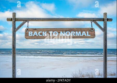 Holzschild mit Text wieder normal gegen blauen Himmel, Meer und Strand mit weißem Sand. Reise- und Urlaubskonzept. Stockfoto