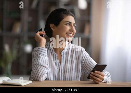 Lächelnde verträumte indische Frau mit Kreditkarte und Smartphone Stockfoto