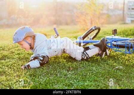 Ein Kind in einem Helm und Schutz fiel von einem Fahrrad auf das Gras und wurde nicht verletzt auf einem Sonniger Tag im Park Stockfoto