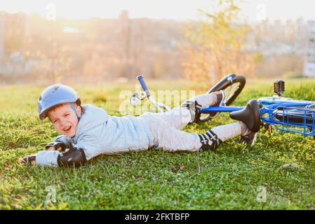 Ein Kind in einem Helm und Schutz fiel von einem Fahrrad auf das Gras und wurde nicht verletzt auf einem Sonniger Tag im Park Stockfoto