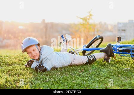 Ein Kind in einem Helm und Schutz fiel von einem Fahrrad auf das Gras und wurde nicht verletzt auf einem Sonniger Tag im Park Stockfoto