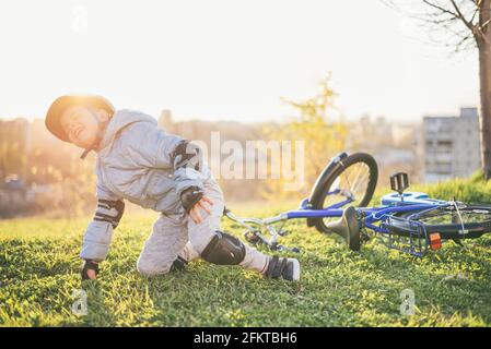 Ein Kind in einem Helm und Schutz fiel von einem Fahrrad auf das Gras und wurde nicht verletzt auf einem Sonniger Tag im Park Stockfoto