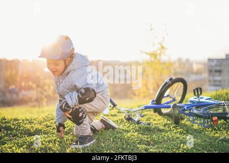 Ein Kind in einem Helm und Schutz fiel von einem Fahrrad auf das Gras und wurde nicht verletzt auf einem Sonniger Tag im Park Stockfoto