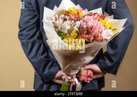 Ein Mann hält einen Blumenstrauß in den Händen und versteckt ihn hinter seinem Rücken mit einem Geschenk mit einer roten Schleife. Überraschung und Geschenke Konzept oder Jubiläum. „Von“ Stockfoto