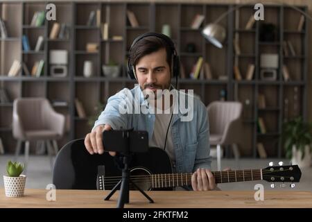 Mann in Kopfhörern hält akustische Gitarre und nimmt Videos auf dem Smartphone auf Stockfoto