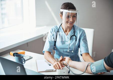 Fröhlicher Arzt in Gesichtsmaske, der den Blutdruck des Patienten überprüft Stockfoto